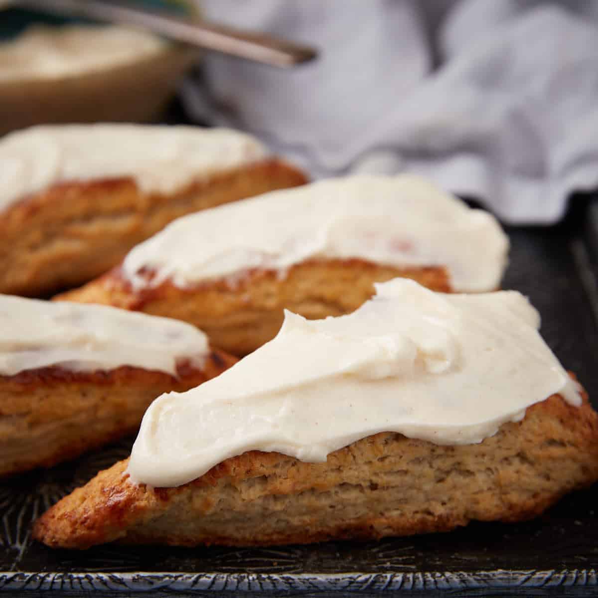 Four triangular scones topped with thick white icing are arranged on a dark tray, with a blurred cloth and a bowl of icing in the background.
