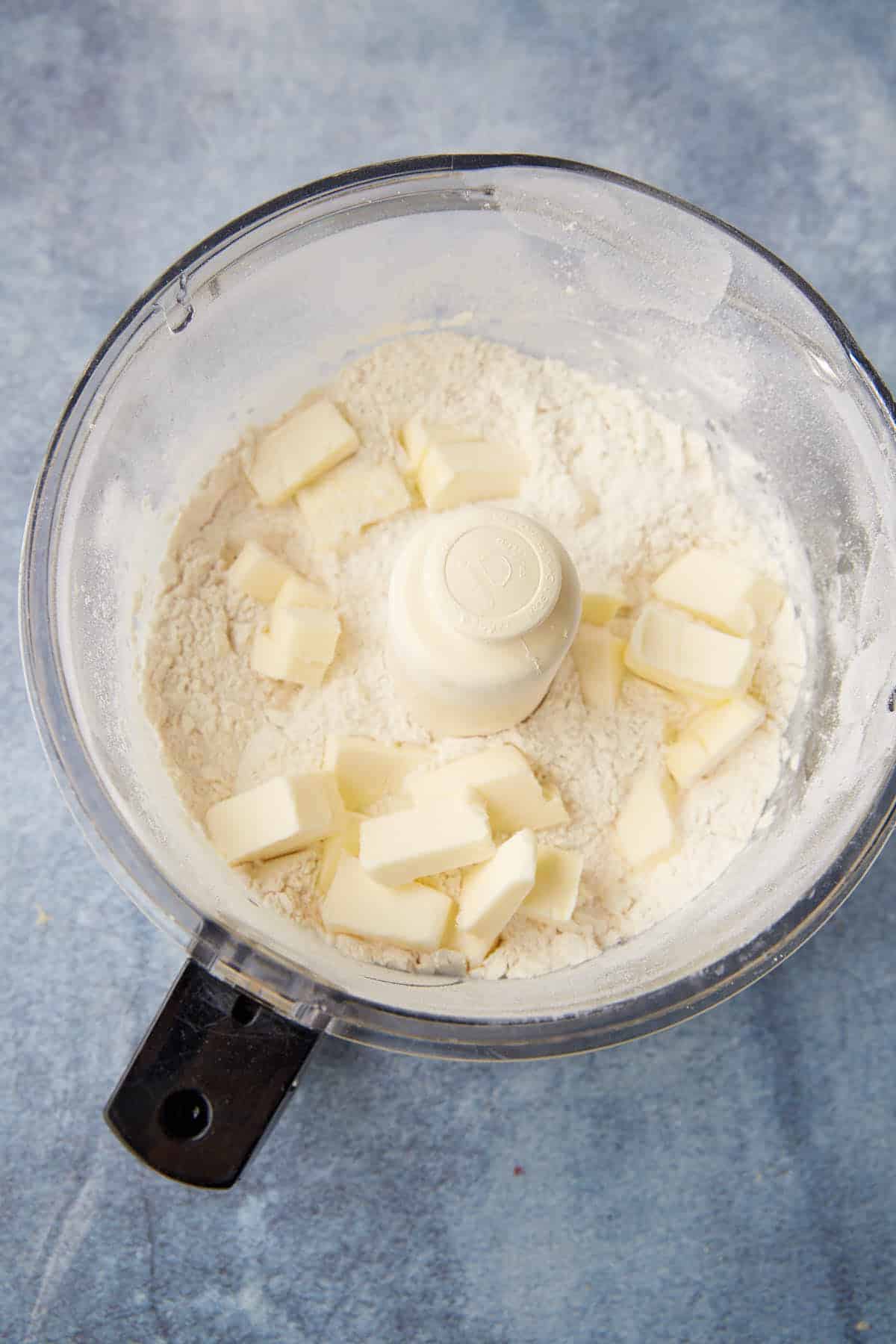 Overhead view of a food processor bowl with flour and small cubes of butter inside, ready to be mixed, on a blue-grey surface.