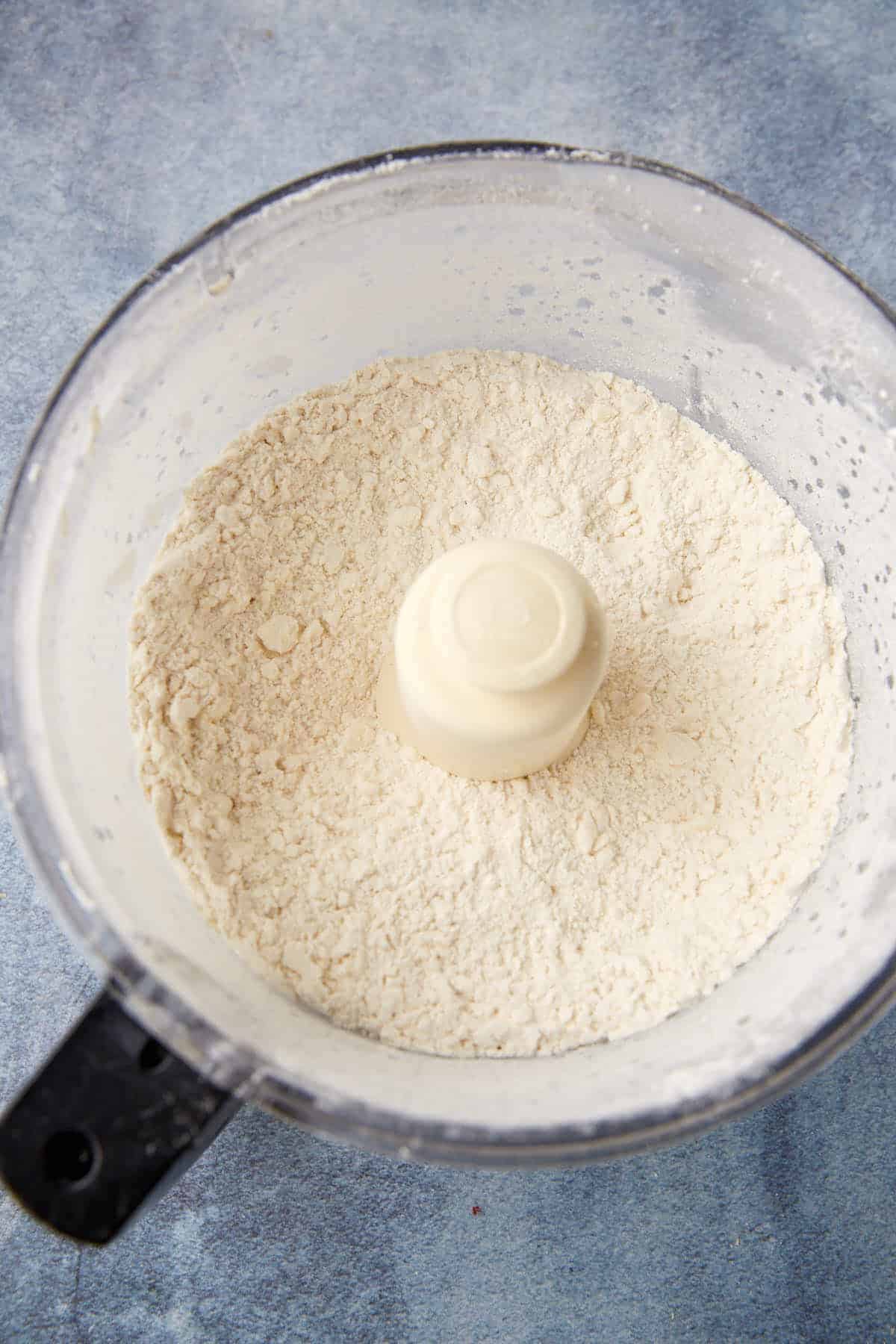 A food processor bowl filled with flour mixture sits on a blue-gray surface, viewed from above. The blade is partially covered by the powdery mixture.