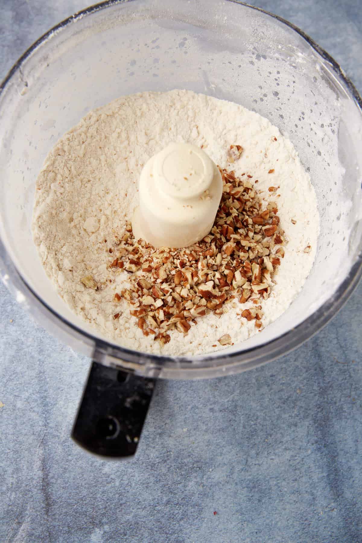A food processor bowl contains flour and chopped pecans on a blue surface, ready for mixing.