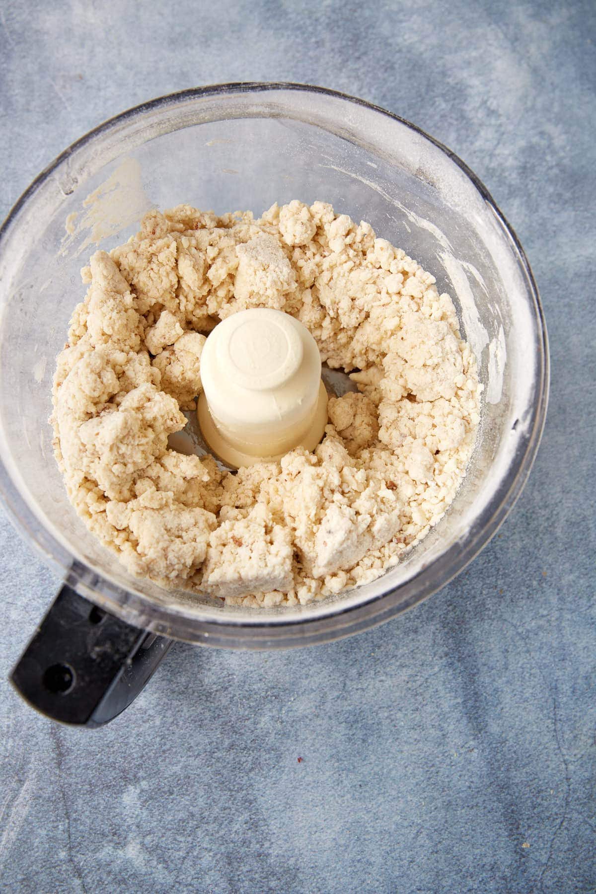 A food processor with coarse, crumbly dough inside sits on a gray countertop, viewed from above.