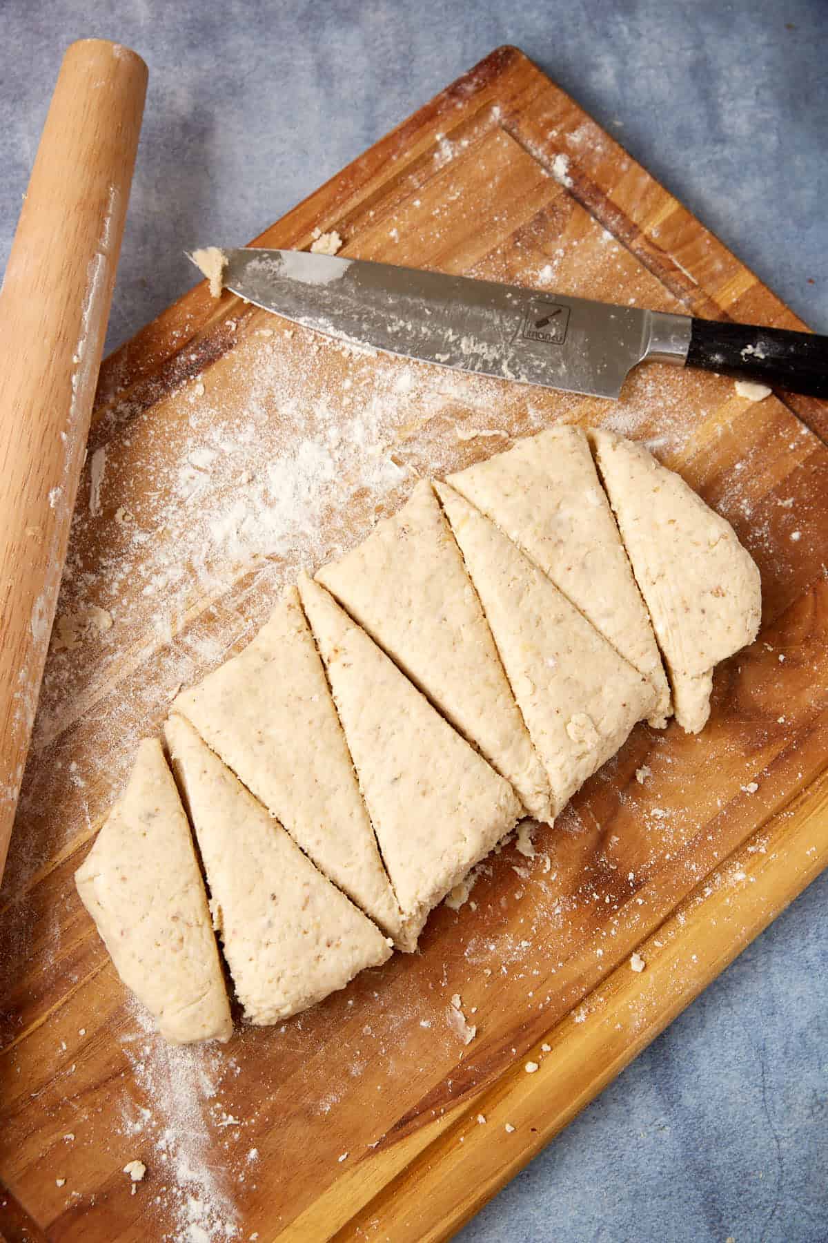 A wooden board holds triangle-shaped pieces of dough, a rolling pin, and a flour-dusted knife, all set against a blue background.