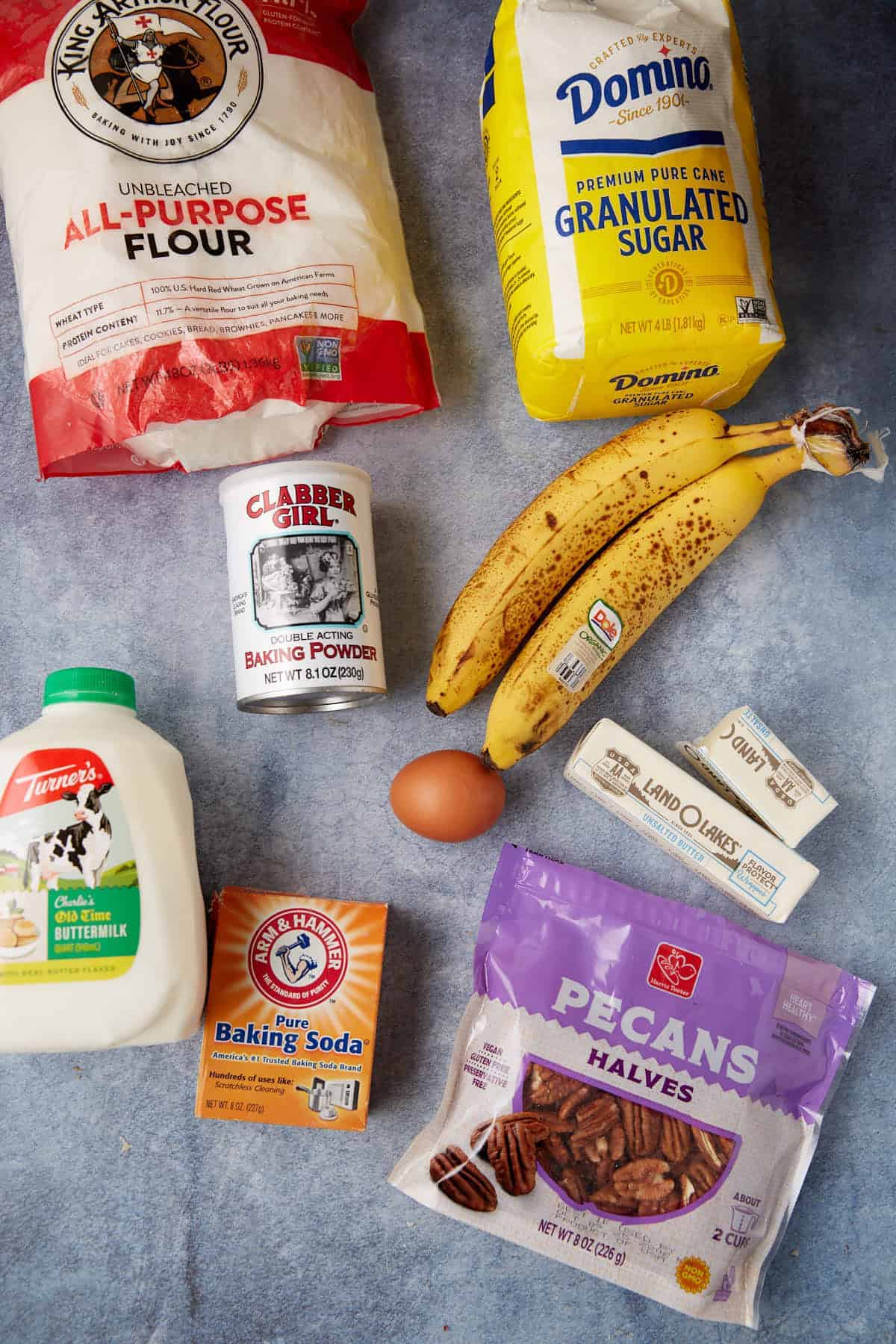 Baking ingredients on a blue surface: flour, sugar, buttermilk, baking powder, baking soda, pecan halves, two bananas, an egg, and two sticks of butter.