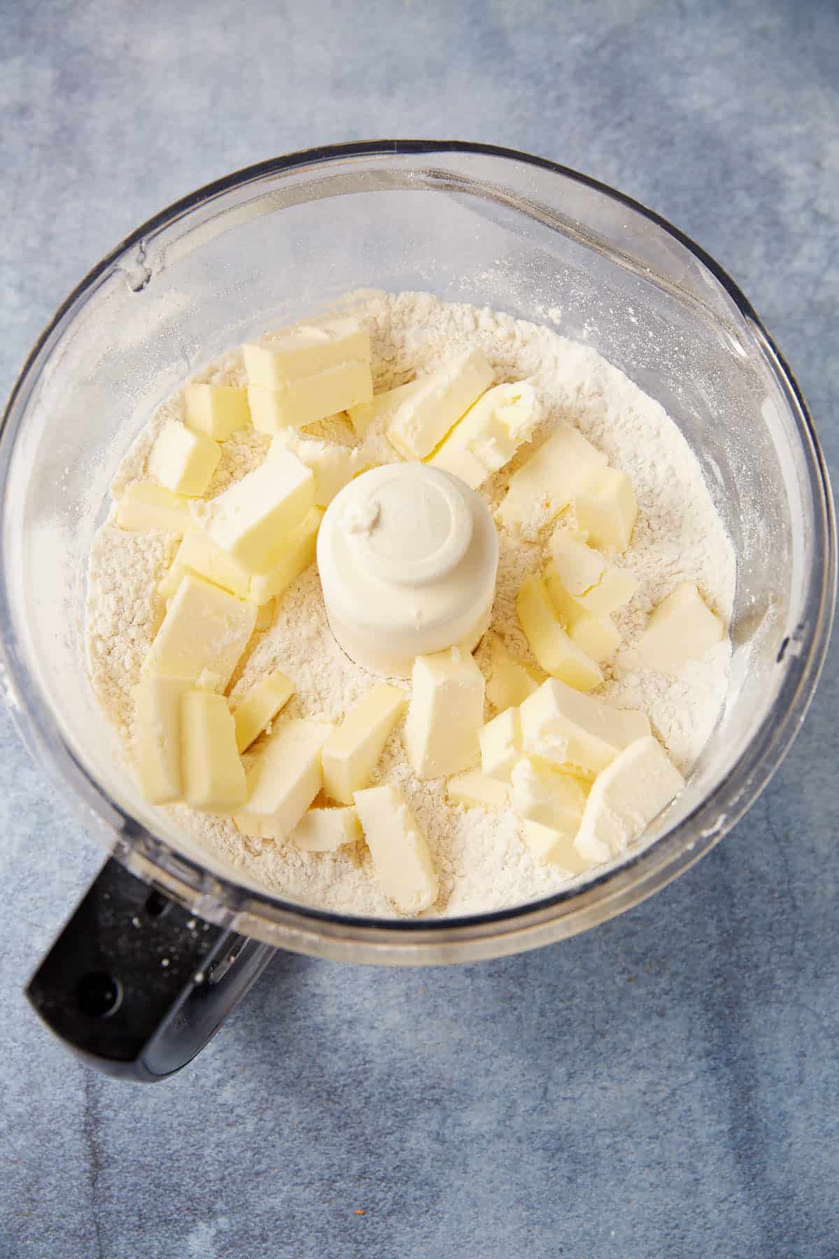 A food processor bowl with flour and cubes of butter inside, ready to be mixed for delicious Chocolate Chip Biscuits on a light blue countertop.