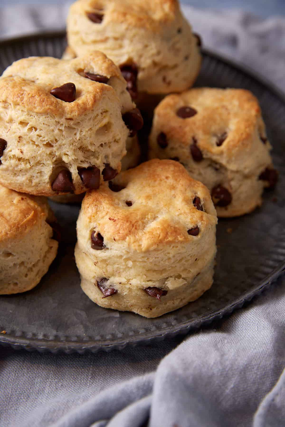 A close-up of chocolate chip scones stacked on a dark plate, with a textured gray cloth underneath. Reminiscent of classic Chocolate Chip Biscuits, the scones are golden brown and fluffy, showing visible chocolate chips.