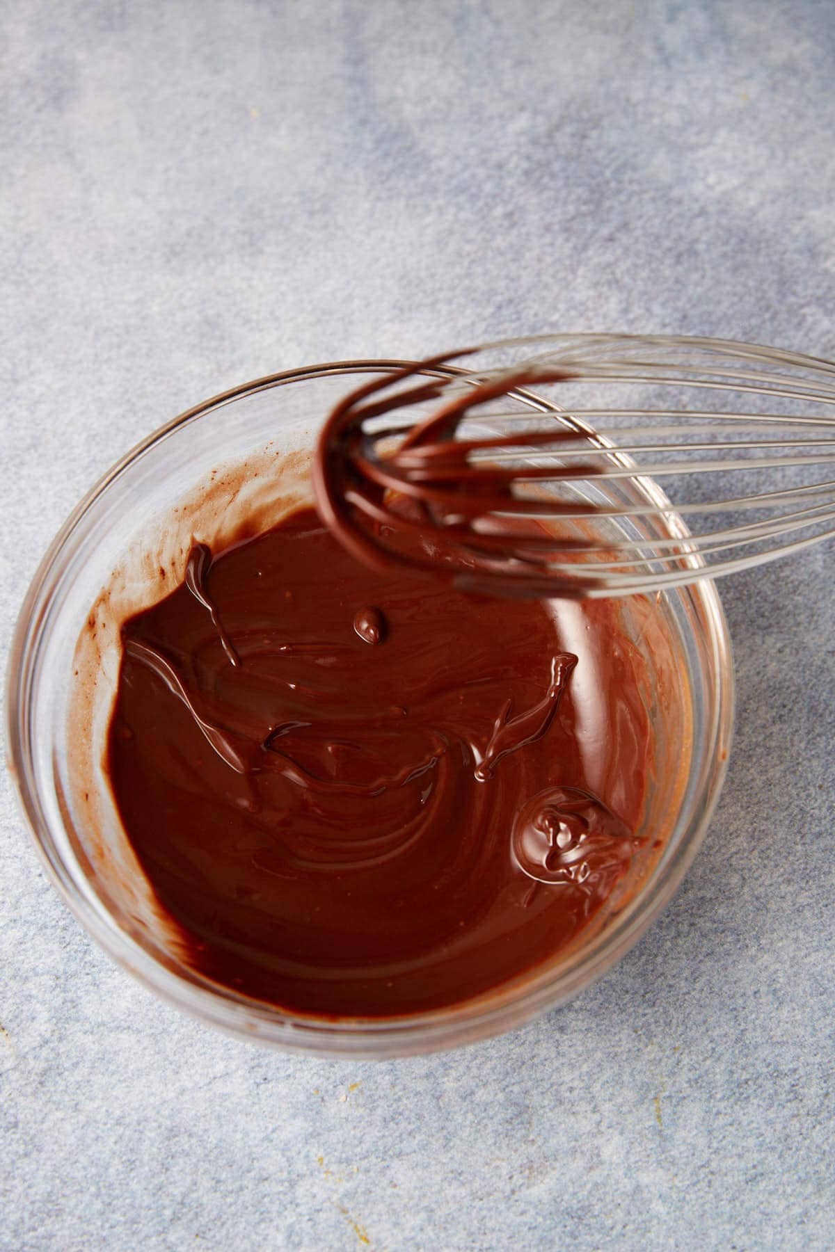 A glass bowl filled with melted chocolate, perfect for making Chocolate Chip Biscuits, is being whisked with a metal whisk and placed on a light gray textured surface.