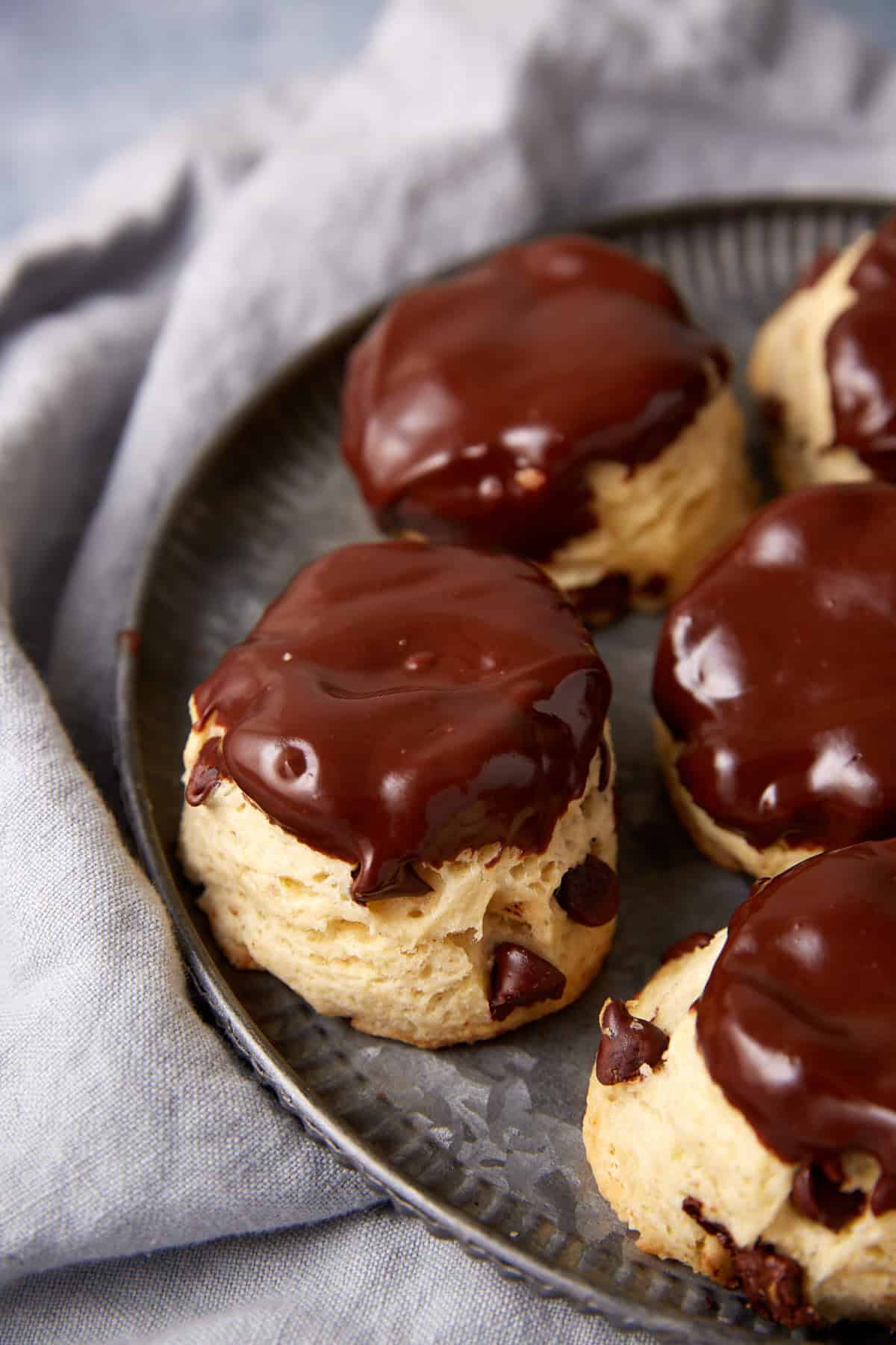 Four chocolate chip scones, reminiscent of classic chocolate chip biscuits, are topped with glossy chocolate icing and arranged on a round metal plate, set on a soft grey cloth.