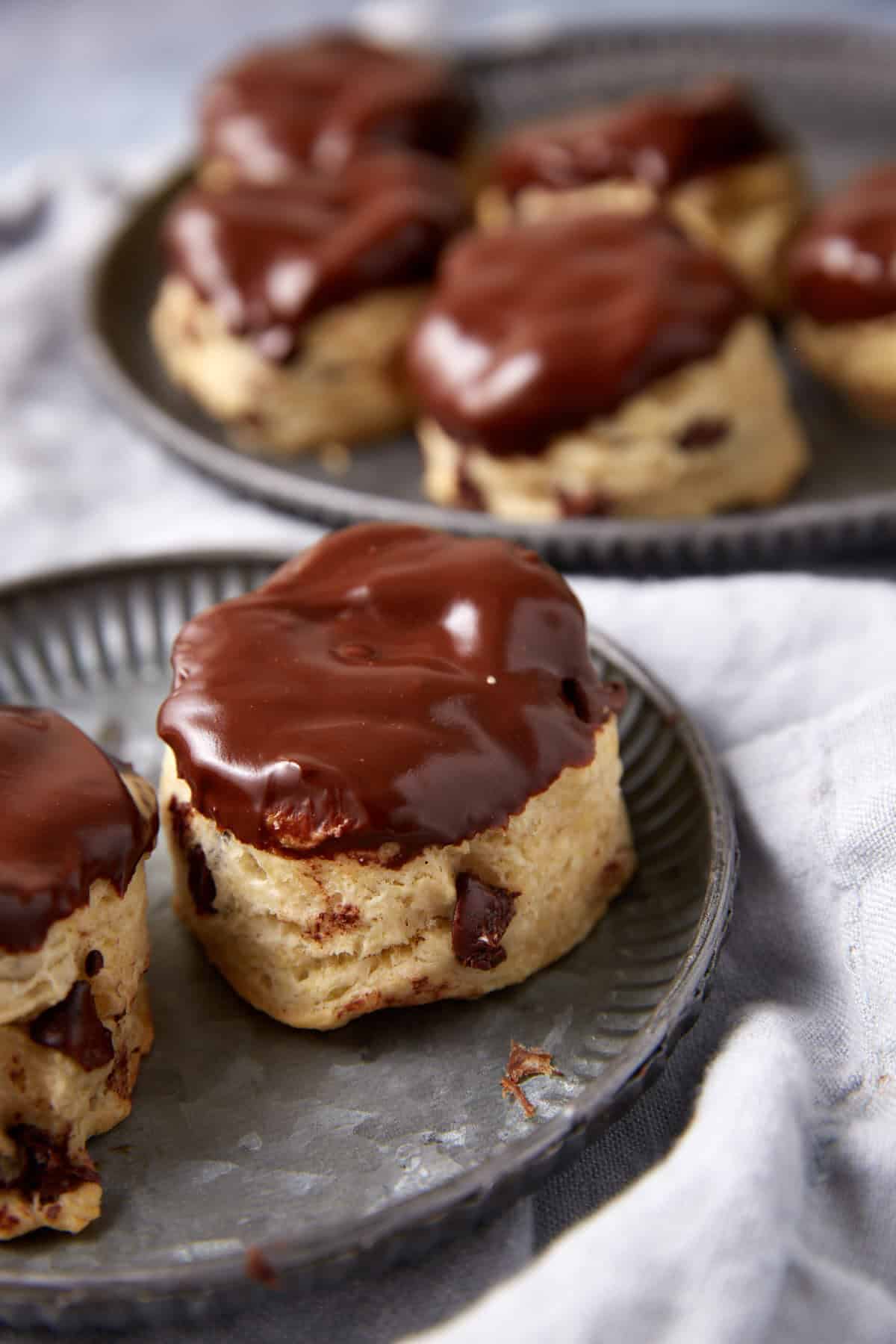 Chocolate Chip Biscuits covered in rich chocolate, with visible chocolate chips peeking through, are arranged on metal plates. The background is softly blurred, and a light cloth is set underneath for an inviting presentation.
