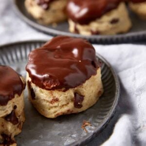 Three chocolate chip scones, reminiscent of classic Chocolate Chip Biscuits, are topped with glossy chocolate glaze and sit on a metal plate, with more scones visible in the background on a gray cloth.