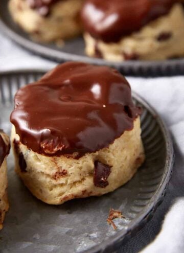 Three chocolate chip scones, reminiscent of classic Chocolate Chip Biscuits, are topped with glossy chocolate glaze and sit on a metal plate, with more scones visible in the background on a gray cloth.