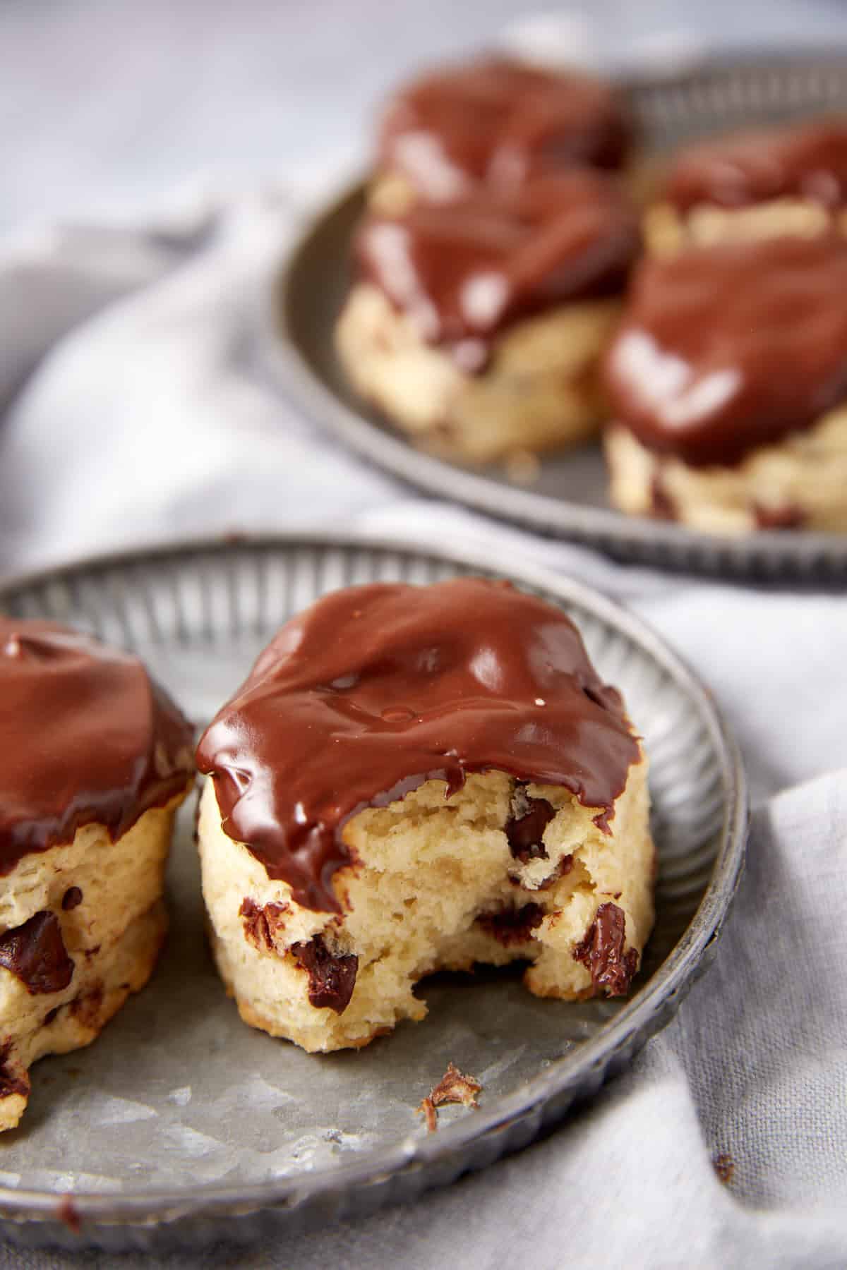 Two chocolate chip scones, reminiscent of classic chocolate chip biscuits, are topped with a rich chocolate glaze and sit on a plate, one with a bite taken out. More scones are visible on a plate in the background.