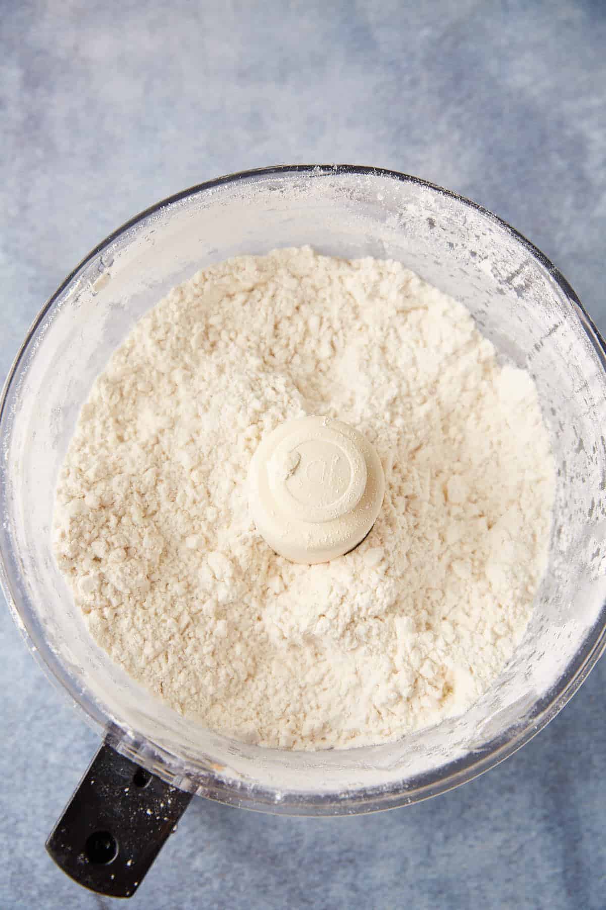 A food processor bowl filled with flour and small pieces of butter, ready to be mixed for making chocolate chip biscuit dough. The background is a light blue surface.