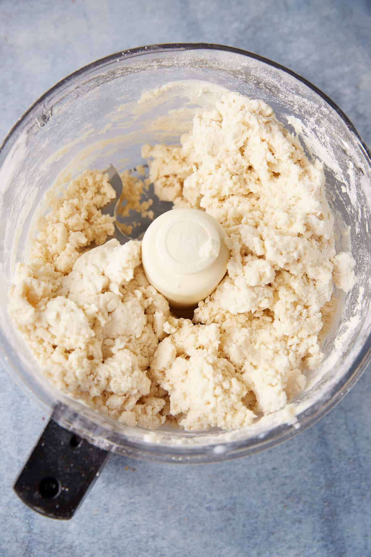 A food processor bowl filled with partially mixed, crumbly chocolate chip biscuit dough, sitting on a light blue surface.