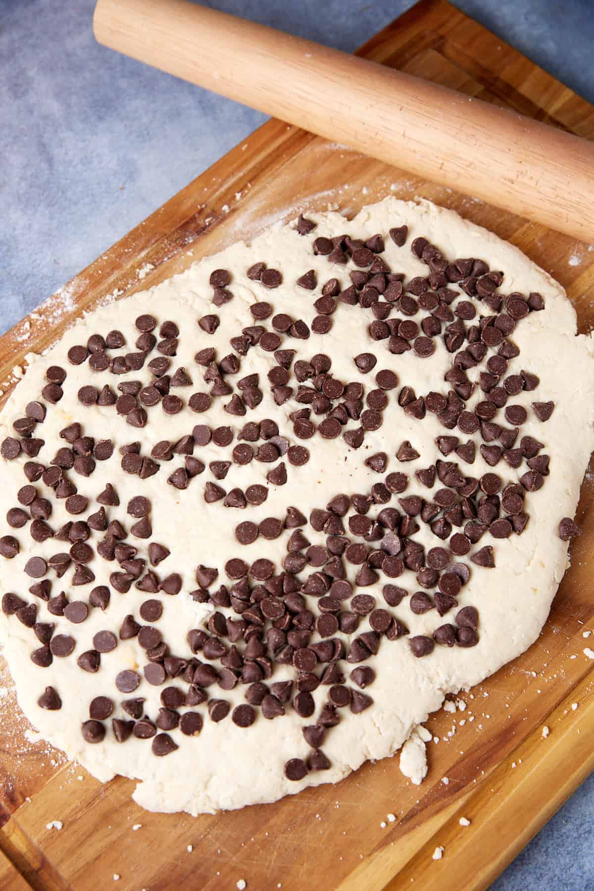Rolled-out dough for Chocolate Chip Biscuits topped with chocolate chips on a wooden cutting board, with a rolling pin nearby.