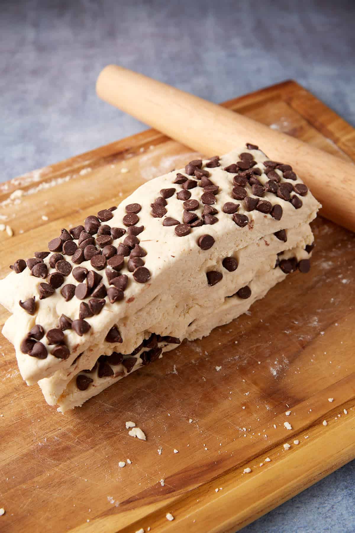 A block of chocolate chip cookie dough, soon to become delicious chocolate chip biscuits, rests on a wooden cutting board next to a rolling pin. Chocolate chips are scattered throughout the dough.