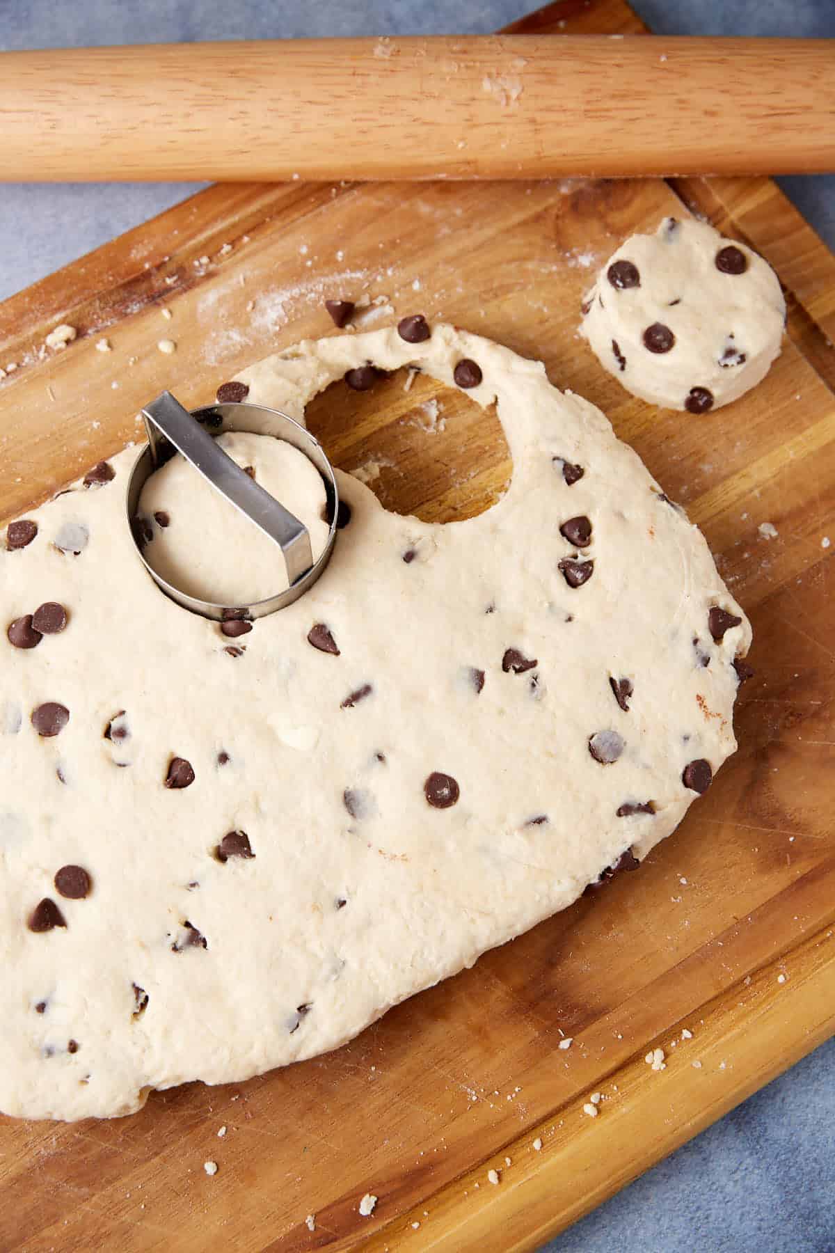 Chocolate chip-studded dough on a wooden board with a round metal biscuit cutter and a rolling pin, ready for baking delicious Chocolate Chip Biscuits.
