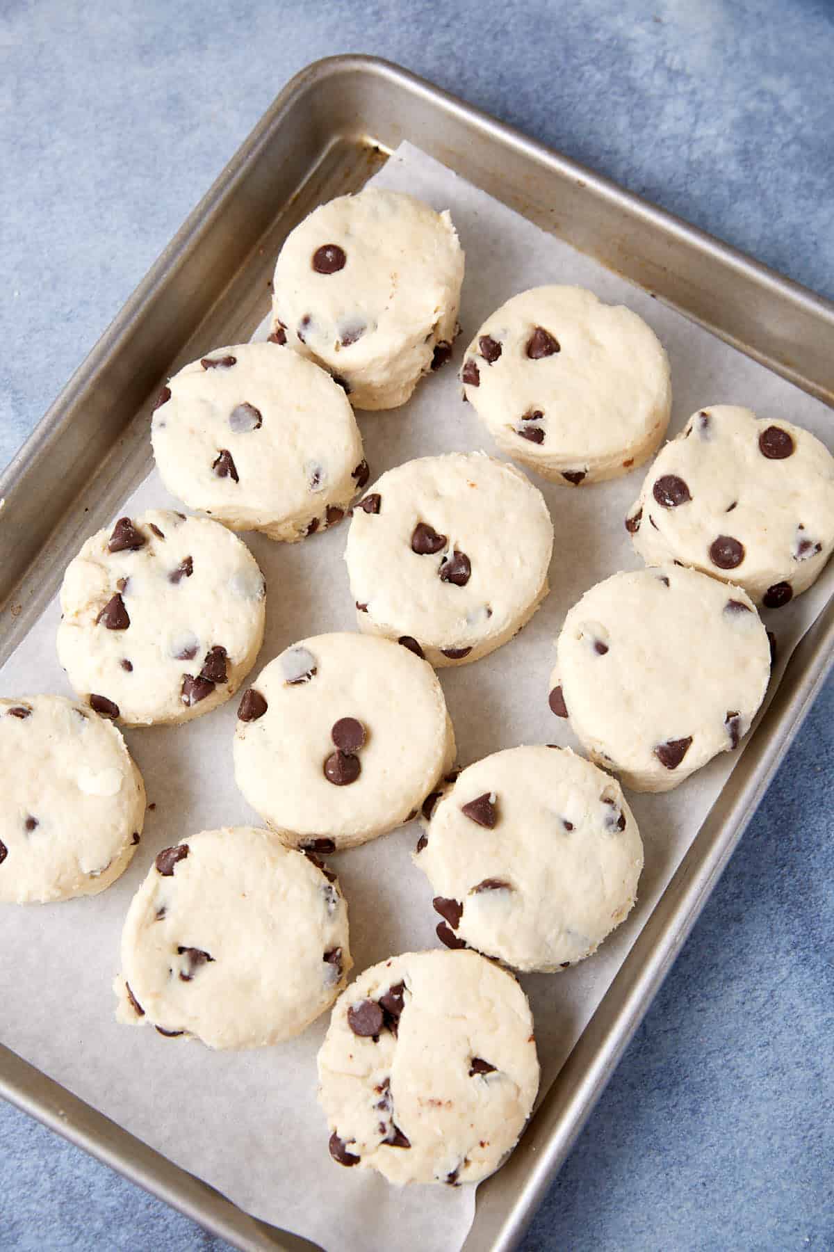 Twelve unbaked chocolate chip scones, reminiscent of classic Chocolate Chip Biscuits, are arranged on a parchment-lined baking sheet, ready to be baked. The background is a gray-blue countertop.