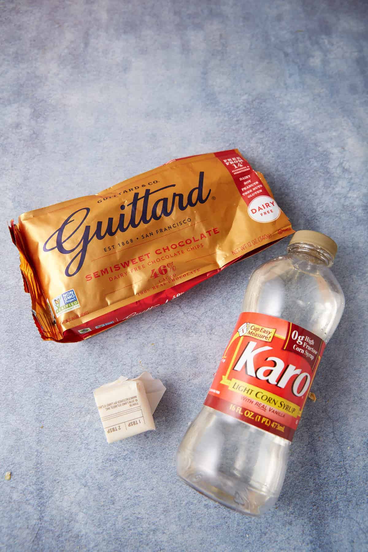 A gold-wrapped Guittard semisweet chocolate bar, an empty Karo light corn syrup bottle, and a stick of unsalted butter on a blue surface—classic ingredients perfect for baking homemade Chocolate Chip Biscuits.