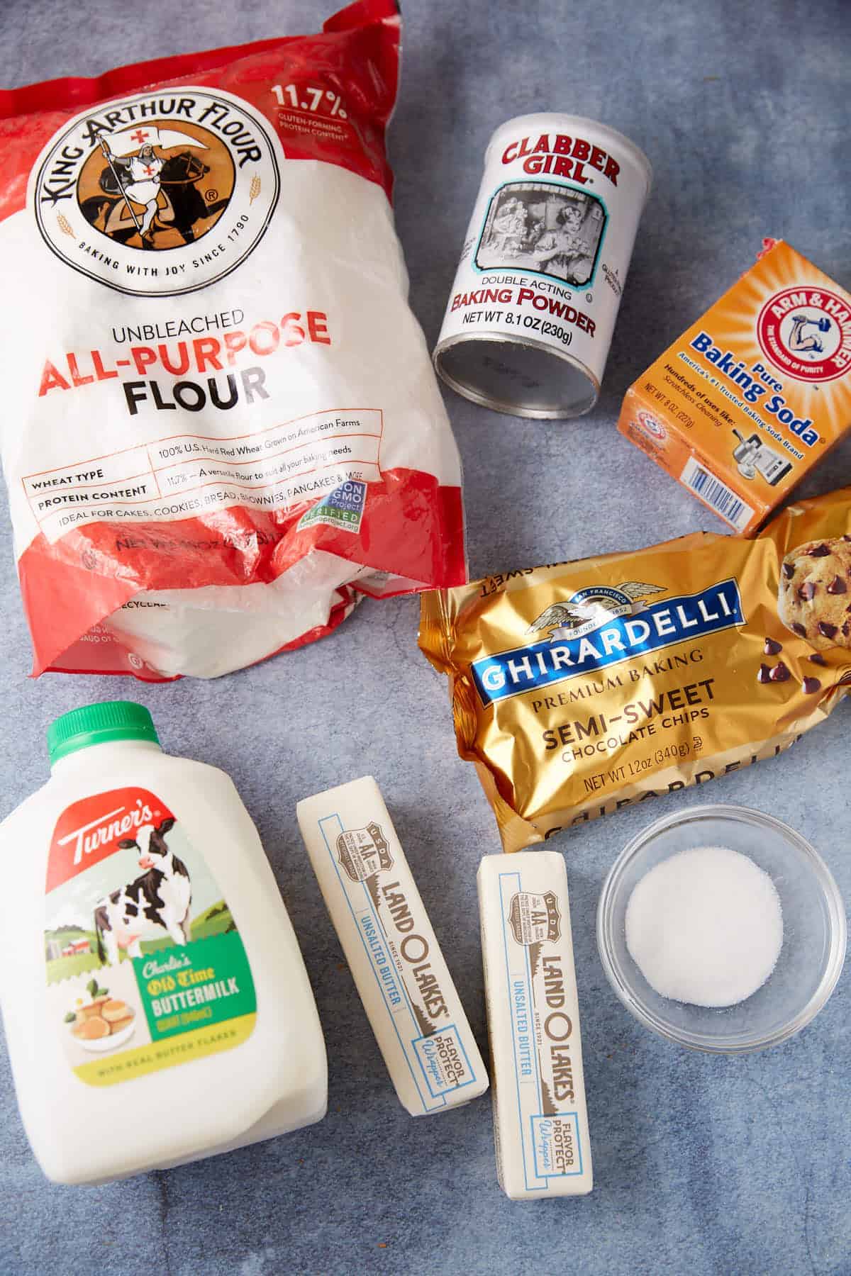 Various baking ingredients on a blue surface, including flour, baking powder, baking soda, chocolate chips, buttermilk, two butter sticks, and a small bowl of sugar—everything you need to whip up delicious Chocolate Chip Biscuits.