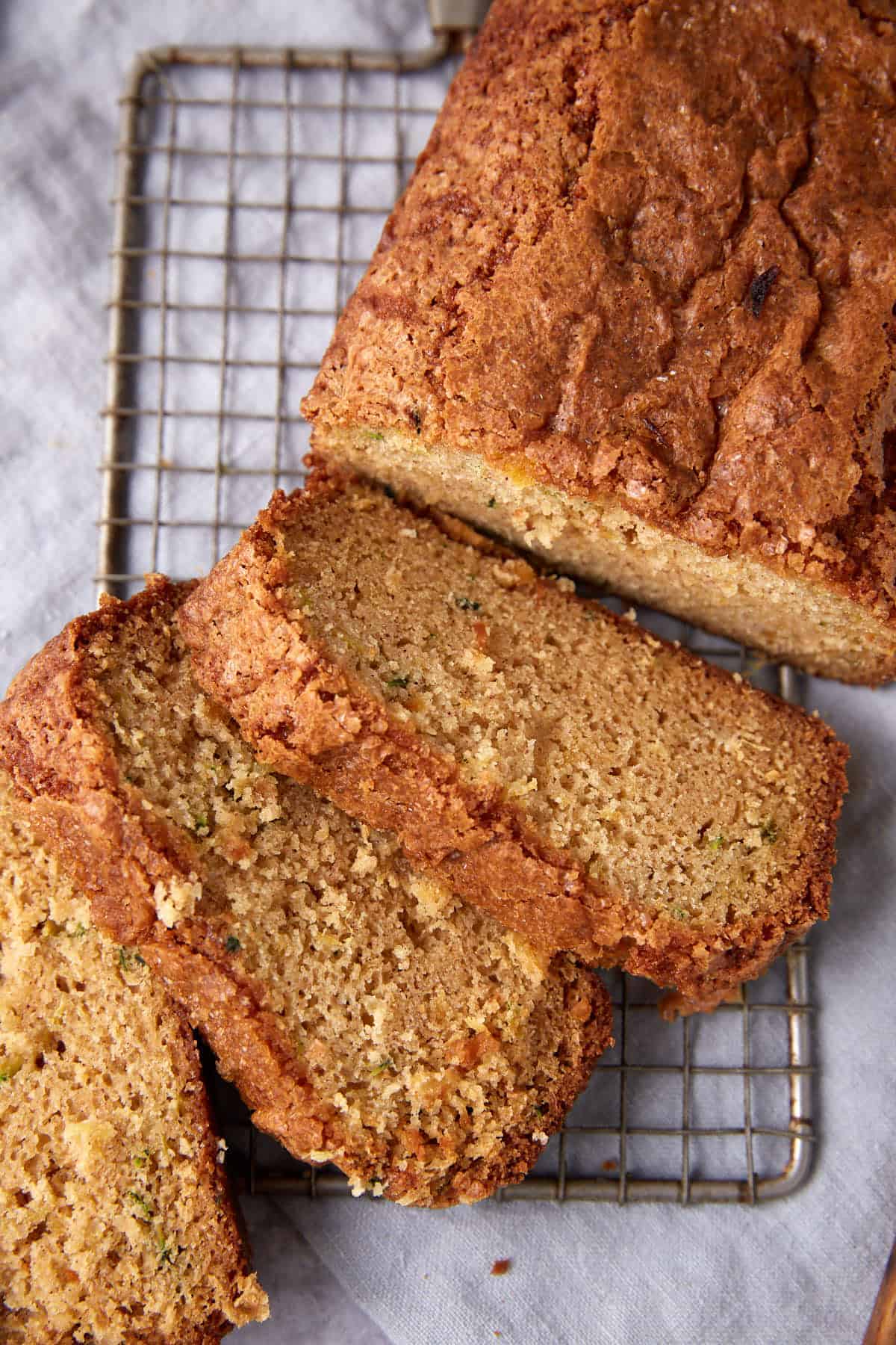 A loaf of zucchini bread with a golden crust is on a cooling rack. Several slices are cut, showing the moist, textured interior on a light cloth background.