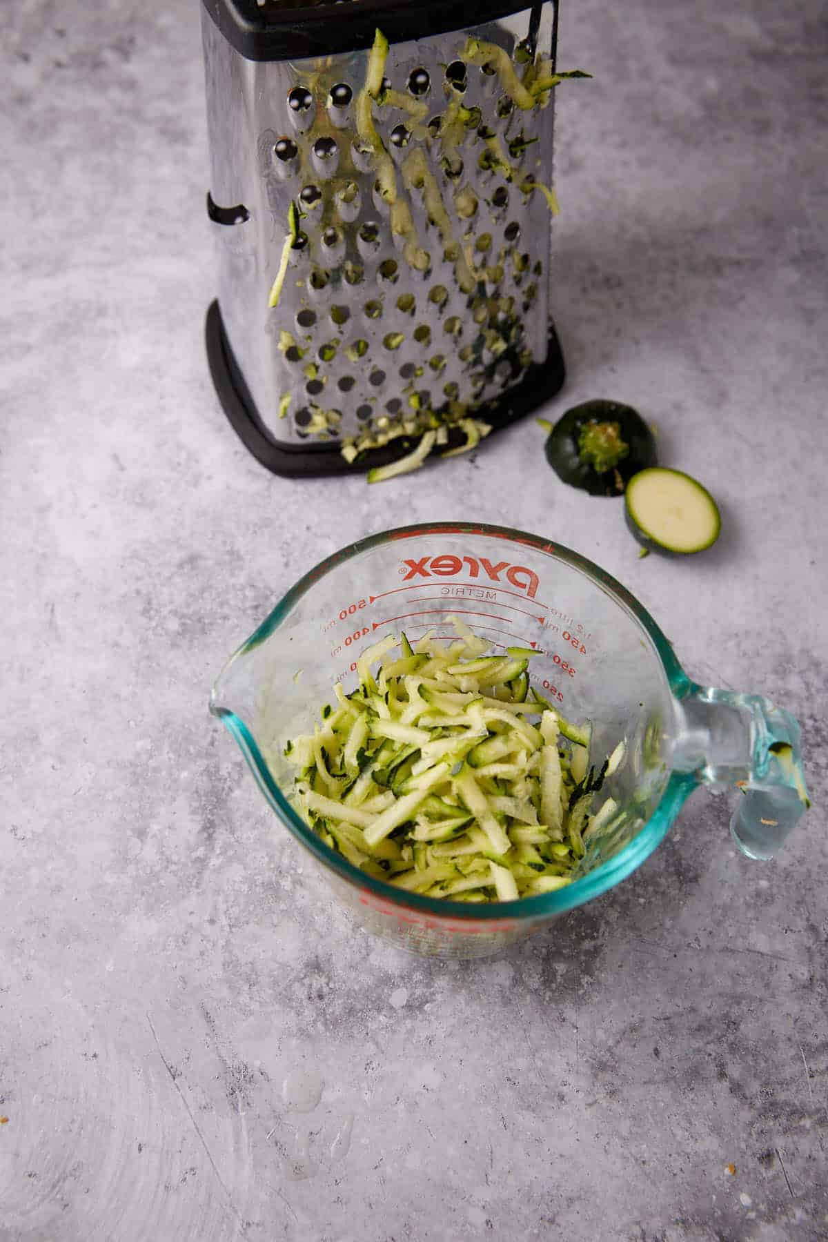 A glass measuring cup filled with shredded zucchini sits on a gray surface, next to a metal box grater and a sliced zucchini end.