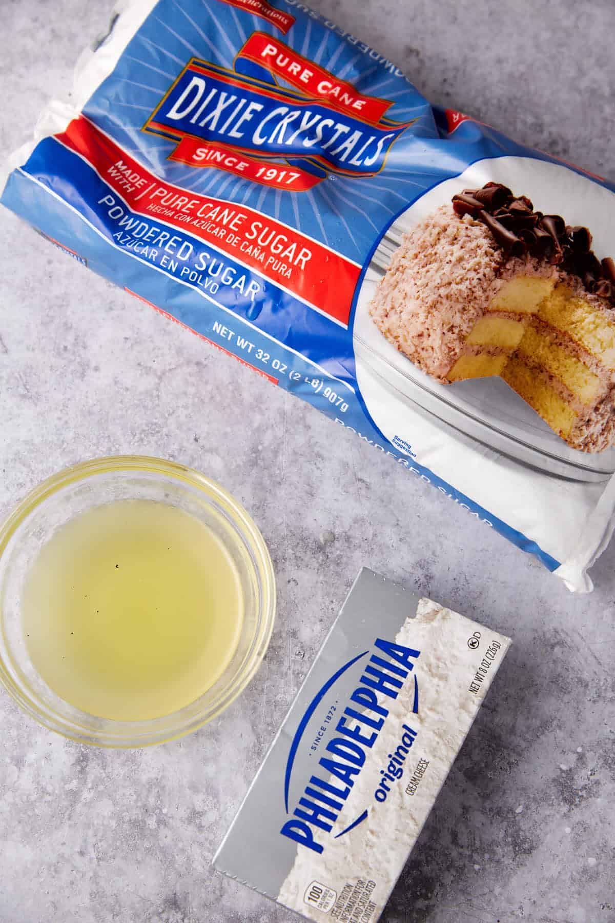 A package of Dixie Crystals powdered sugar, a bowl of egg white, and a box of Philadelphia cream cheese on a gray marble surface.