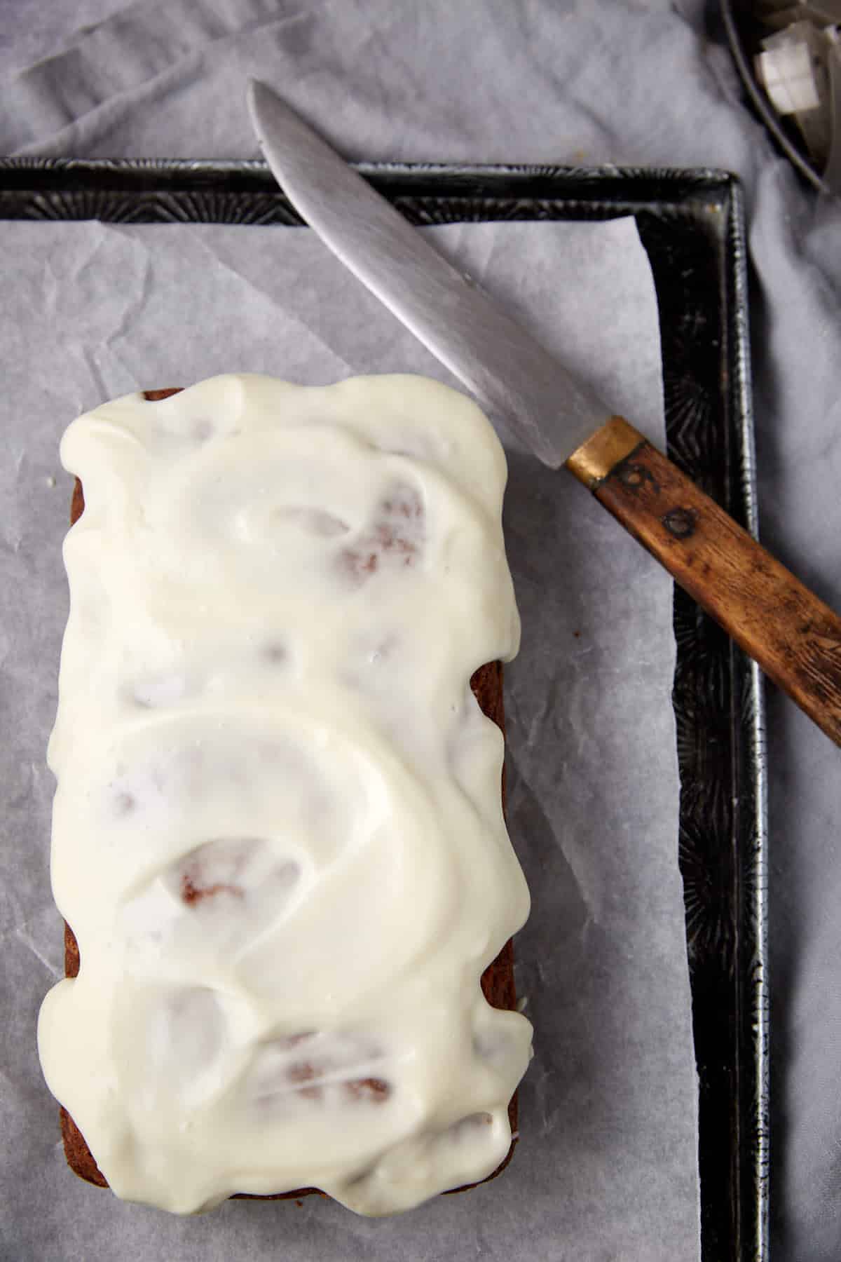 A loaf of frosted bread or cake sits on parchment-lined tray next to a knife with a wooden handle, on a gray fabric background.