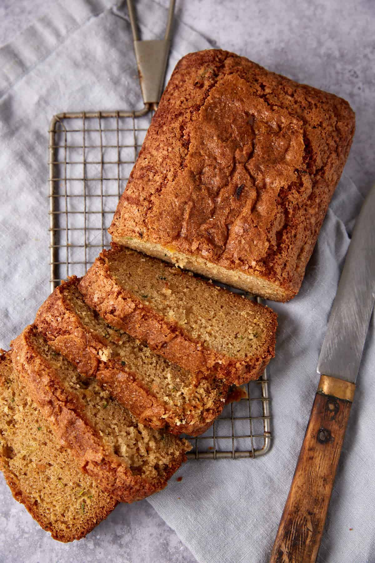A loaf of zucchini bread on a cooling rack, partially sliced, with a serrated knife beside it on a gray cloth.