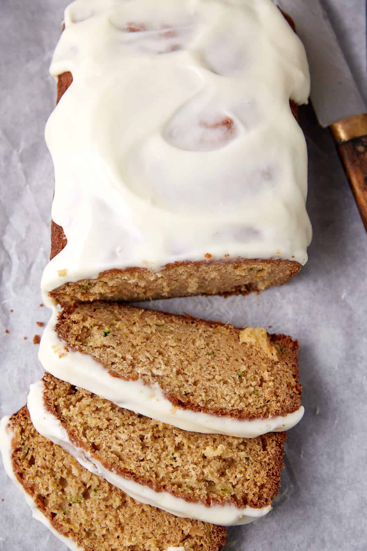 A loaf of zucchini bread with white icing, partially sliced, sits on parchment paper next to a knife with a wooden handle.