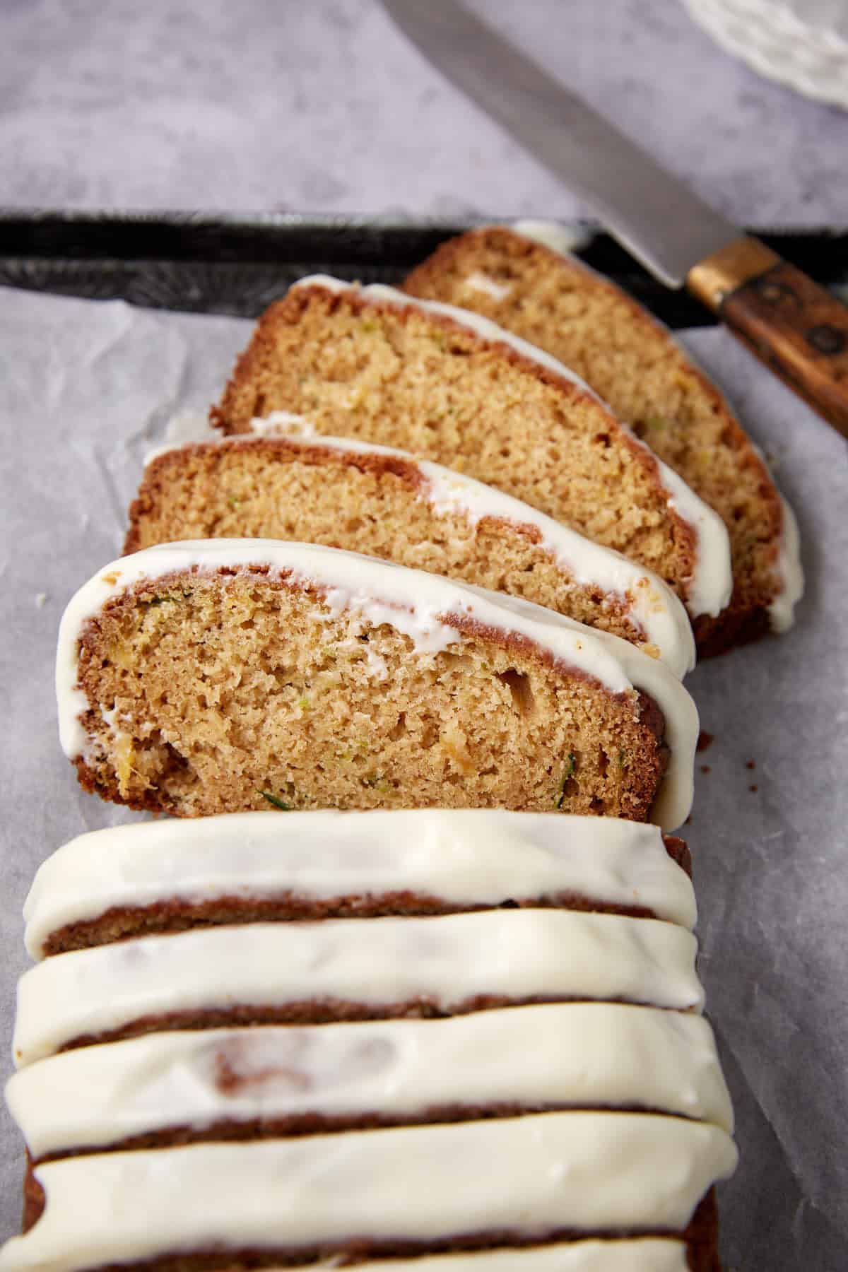 Five slices of zucchini bread with white icing are arranged on parchment paper, with a knife placed nearby.