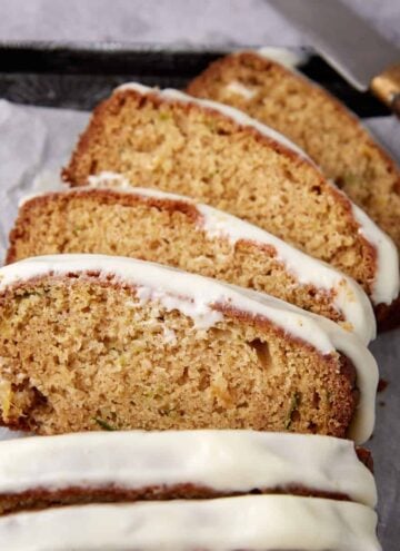 Four slices of zucchini bread with white icing are arranged on parchment paper. A knife with a wooden handle rests nearby.