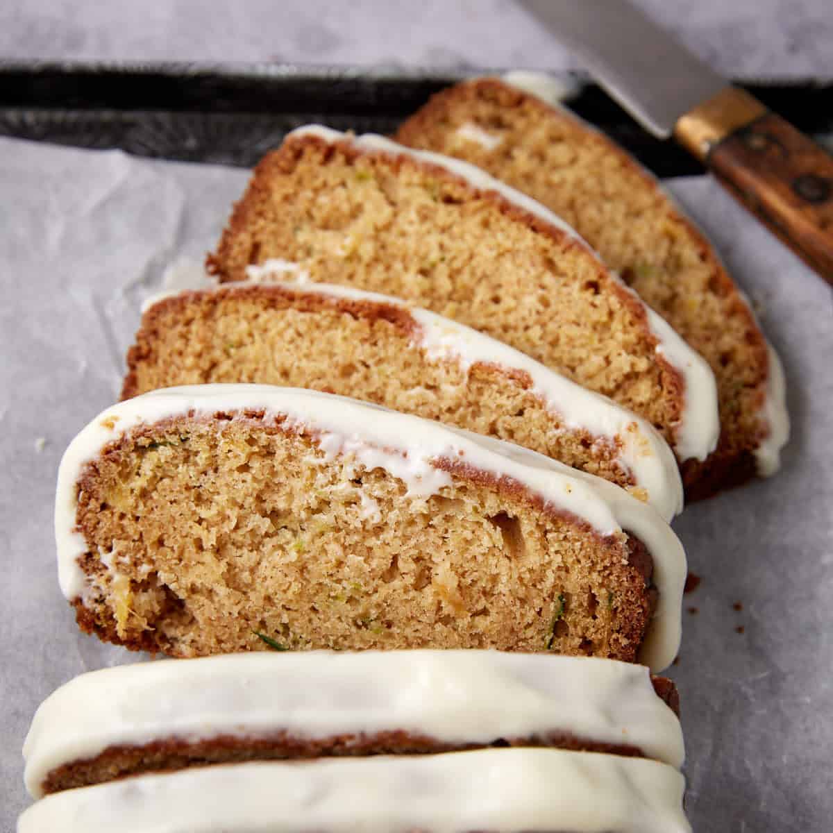 Four slices of zucchini bread with white icing are arranged on parchment paper. A knife with a wooden handle rests nearby.