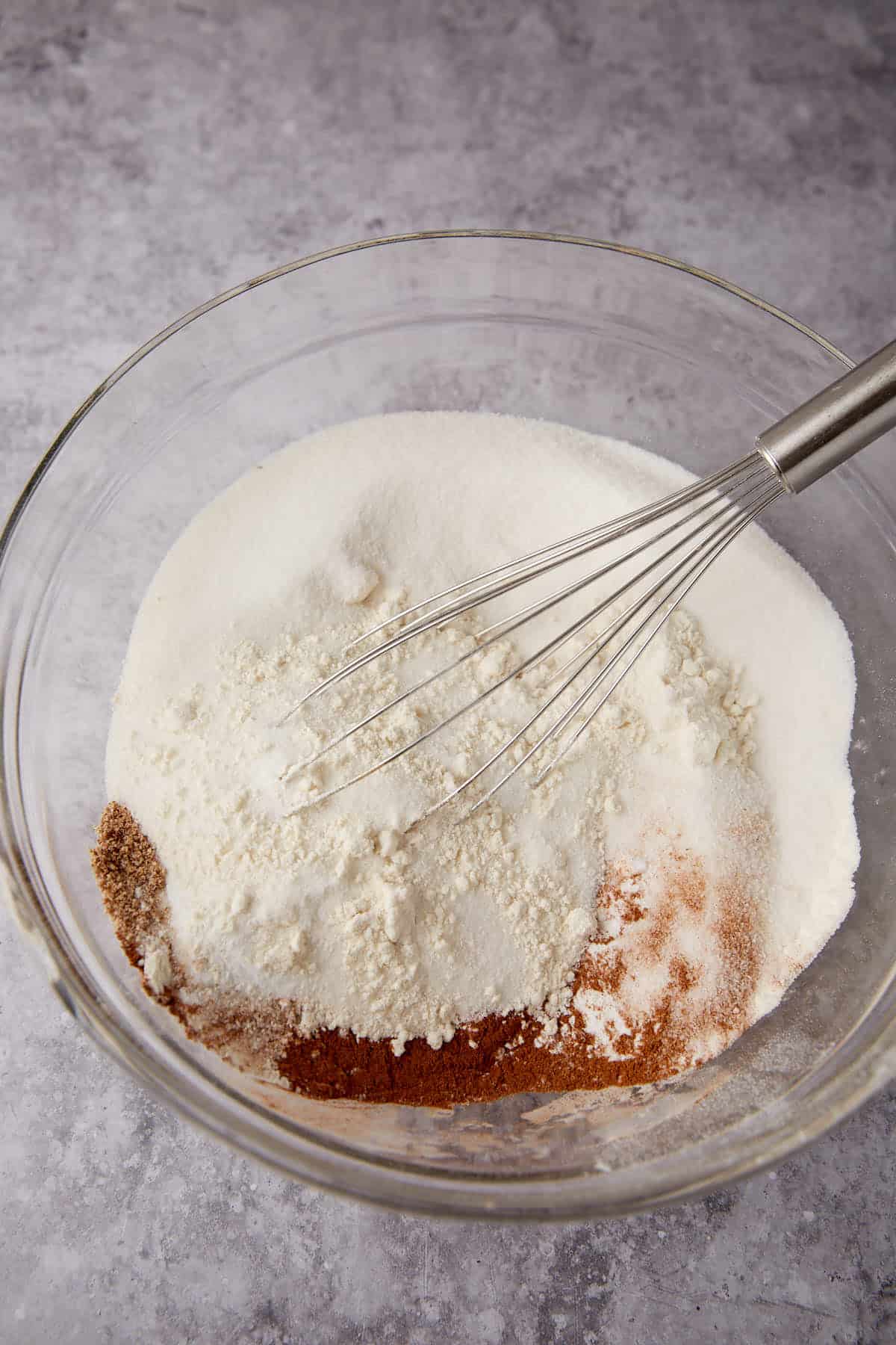 A glass bowl filled with various dry baking ingredients and a metal whisk, ready to be mixed on a gray countertop.