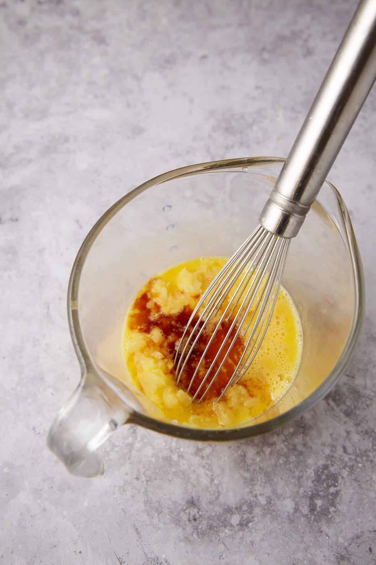 A metal whisk sits in a glass measuring cup containing whisked eggs, mashed bananas, and vanilla extract on a light gray countertop.