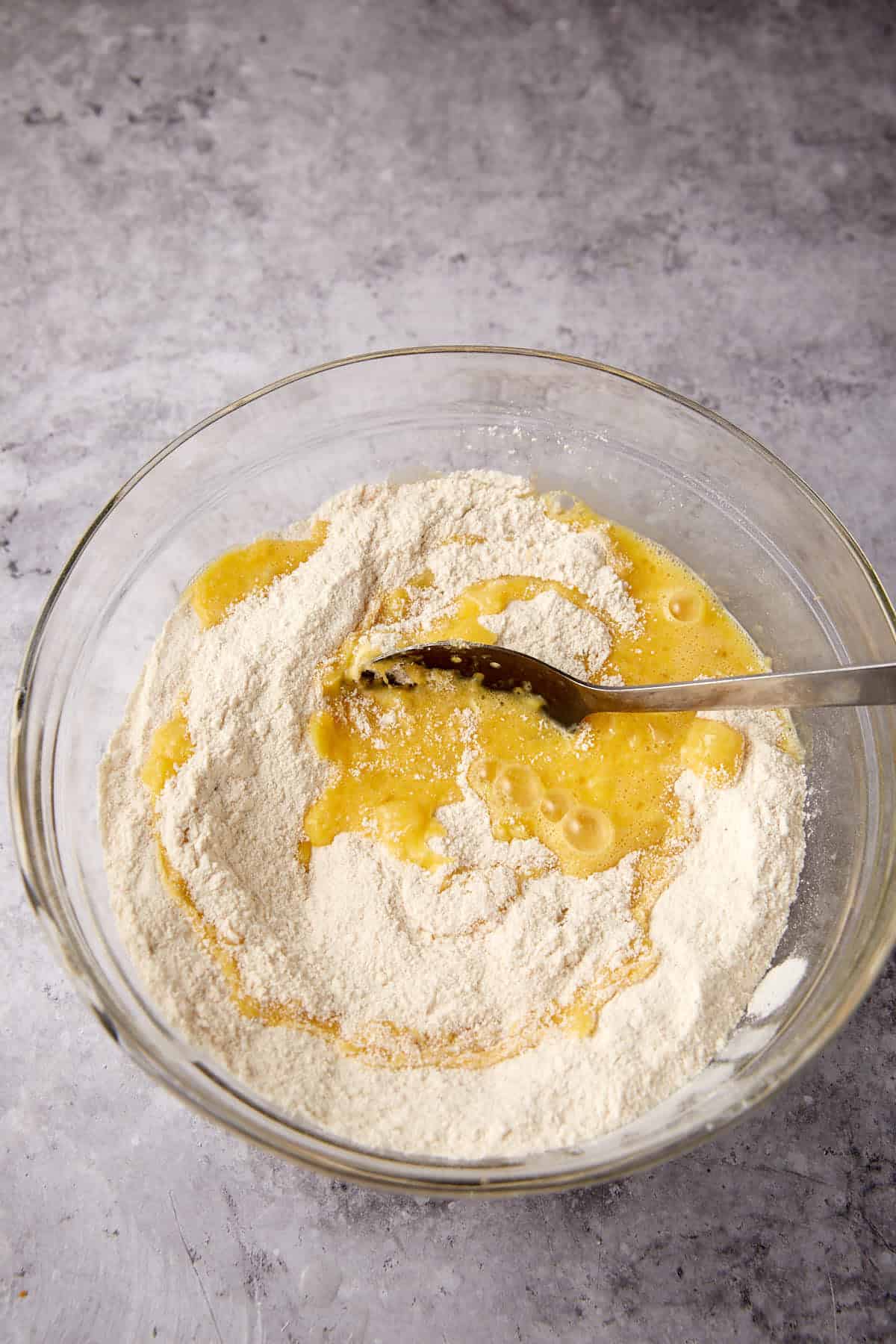 A glass bowl with flour and beaten eggs being mixed with a wooden spoon on a gray countertop.