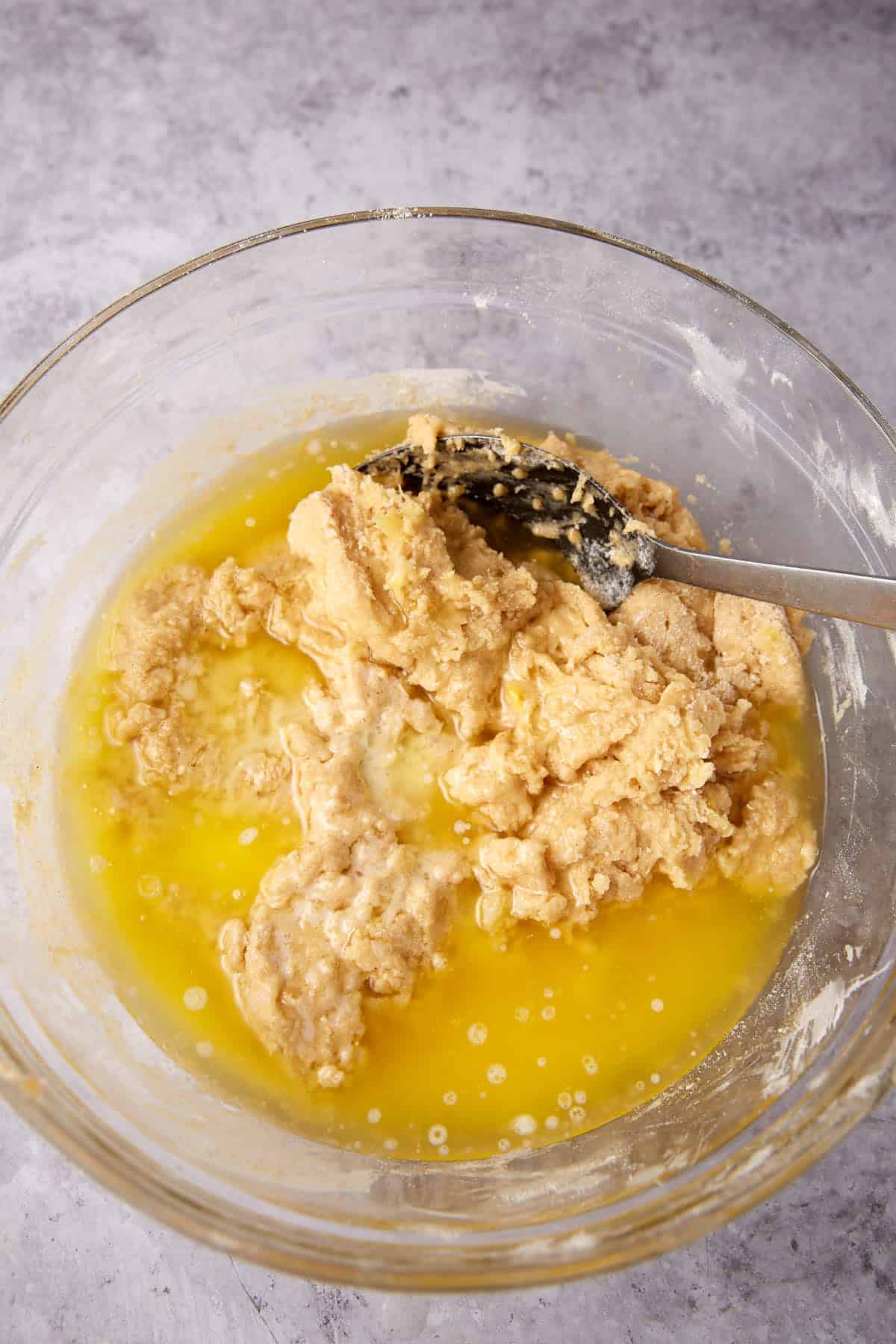 A glass bowl contains partially mixed dough with melted butter pooling around it. A metal spoon rests in the mixture, and the bowl sits on a gray countertop.