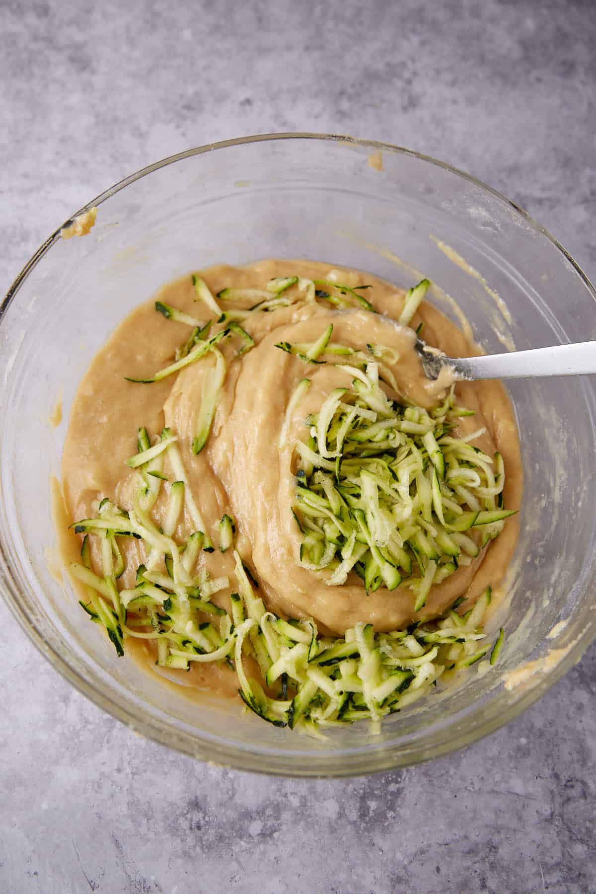 A glass bowl with light brown batter and shredded zucchini, partially mixed with a spoon, on a light gray countertop.