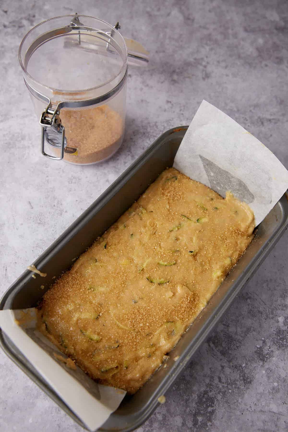A loaf pan lined with parchment paper holds unbaked zucchini bread batter, sprinkled with brown sugar. A container of brown sugar sits nearby on a gray countertop.