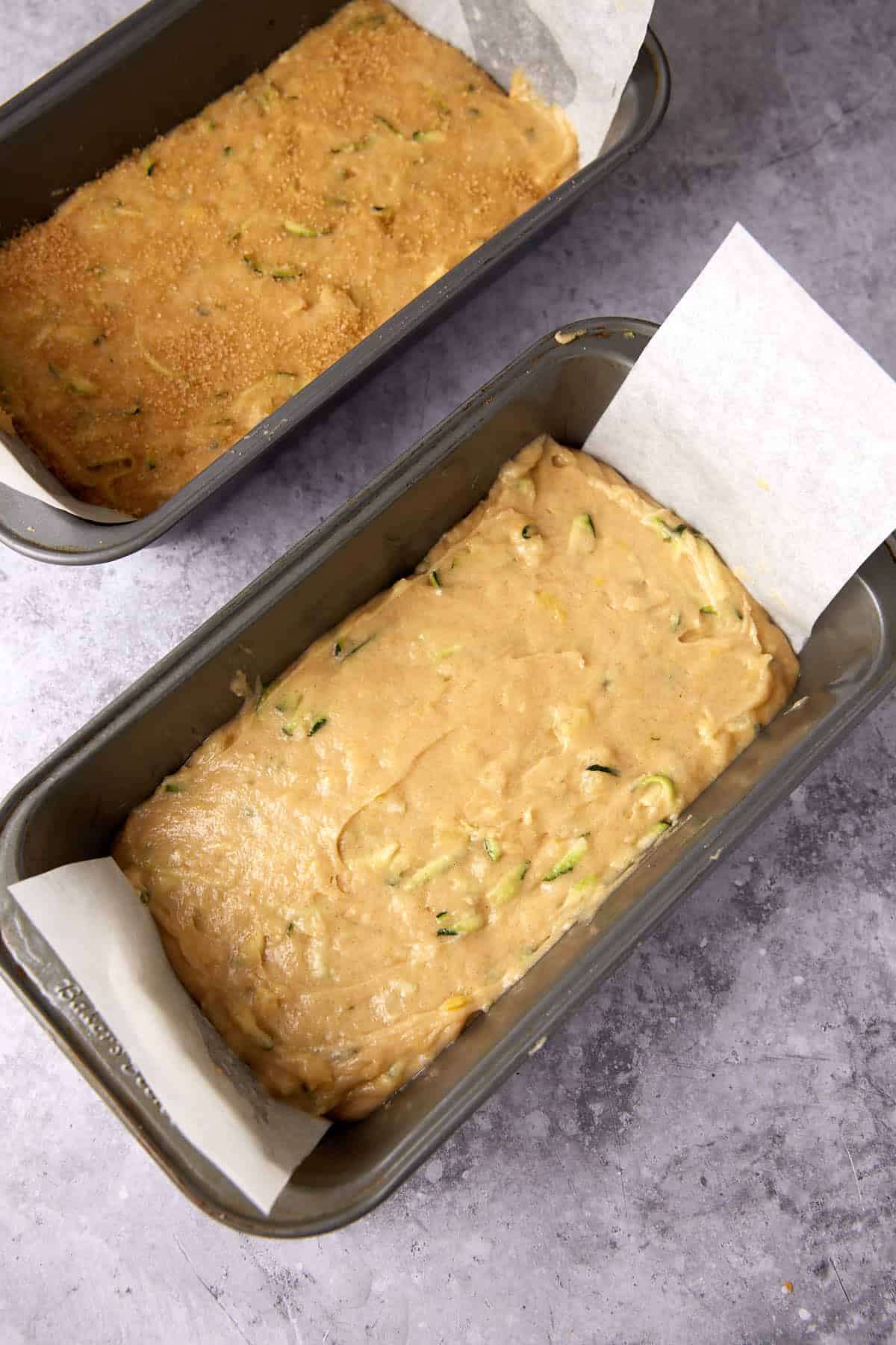 Two metal loaf pans filled with unbaked batter, one with visible shredded zucchini, both lined with parchment paper, placed on a gray textured surface.