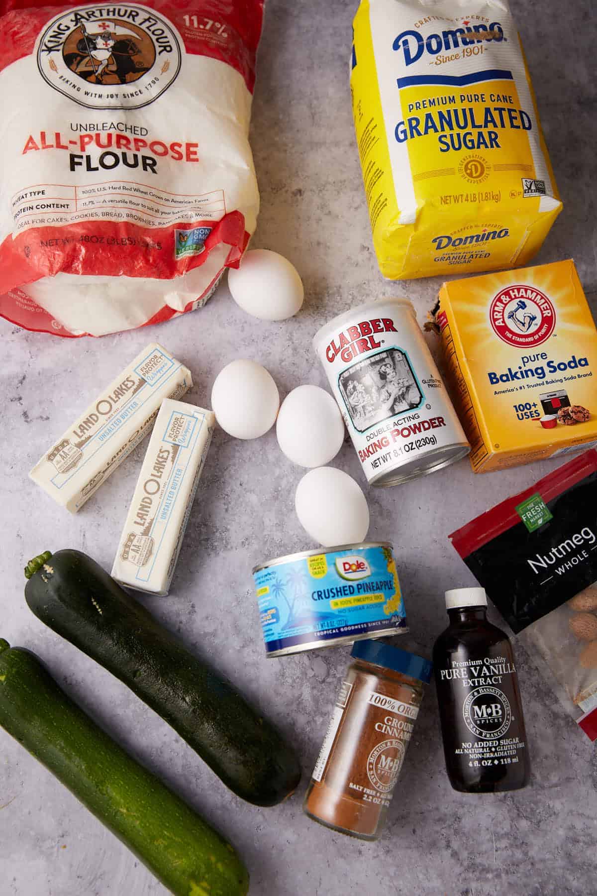 Baking ingredients on a countertop: flour, sugar, eggs, butter, zucchini, canned crushed pineapple, baking powder, baking soda, vanilla extract, ground nutmeg, cinnamon, and salt.