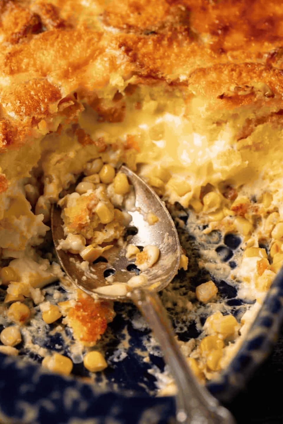 A slotted silver spoon scoops a serving of Southern comfort food, a creamy corn casserole, from a dark blue baking dish.